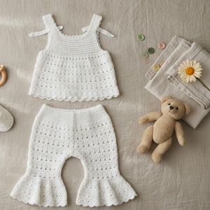 May include: A white crochet two-piece outfit consisting of a sleeveless top with tie-shoulder straps and matching flared trousers. The set features a delicate openwork pattern and scalloped edges, accompanied by white booties, a wooden ring, a teddy bear, and a flower.