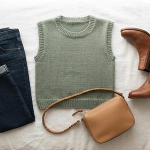 May include: A sage green knit sweater vest with a crew neckline, dark blue denim jeans with cuffed hems, a pair of tan leather Chelsea boots with elastic side panels, and a tan leather shoulder bag featuring a braided strap and gold hardware.