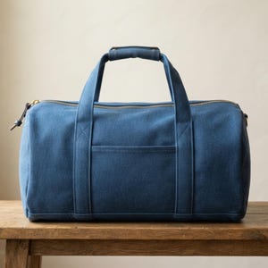 May include: A solid blue duffel bag featuring a horizontal rectangular front pocket, two sturdy top handles with a reinforced grip, and a metallic zipper closure that runs across the top. The bag has a structured cylindrical shape.