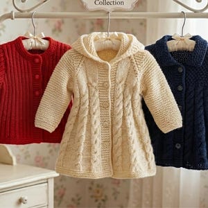 Puede incluir: Tres cárdigans de punto para niños en rojo, crema y azul marino cuelgan de un perchero blanco. El cárdigan rojo tiene una textura acanalada, el crema tiene un diseño con capucha y trenzado, y el azul marino tiene cuello.
