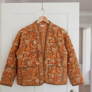 Puede incluir: Chaqueta acolchada con estampado floral en tonos naranja, marrón y crema. La chaqueta, de frente abierto, tiene dos bolsillos y está colgada de una percha de madera. La chaqueta está colgada de una puerta.