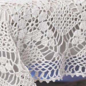 May include: A white crocheted tablecloth featuring a repeating floral motif pattern. The textile is crafted with intricate openwork stitches, creating a delicate lace-like appearance with a scalloped edge along the bottom border of the fabric.