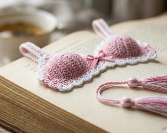May include: A crochet bookmark shaped like a brassiere, featuring two dusty pink cups with white scalloped edges and a small pink ribbon bow in the center. Two braided cords extend from the side, each ending in a pink and white yarn tassel.