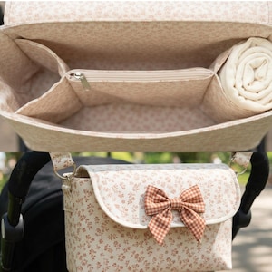 Könnte beinhalten: Eine beige Kinderwagen-Organizer-Tasche mit kleinem braunem Blumenmuster und einer braun-weiß karierten Schleife auf der Frontklappe. Das Innere ist mit demselben Blumenstoff ausgekleidet und enthält ein Reißverschlussfach sowie Seitenfächer.