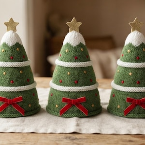 May include: Three cone-shaped knitted decorations resembling Christmas trees. Each green tree features white knitted snow at the top, a gold glitter star topper, white horizontal bands, small red and gold embroidered dots, and a red velvet bow.