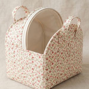 May include: A fabric storage basket featuring a cream background with a small-scale pink and red floral print. The basket has a structured, semi-circular shape with two matching fabric handles and an open top revealing a plain cream interior lining.