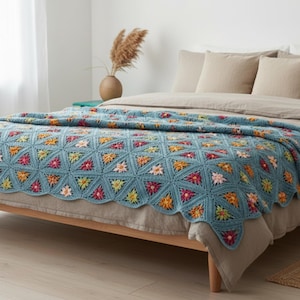 Pode incluir: Uma manta de crochê com um padrão repetitivo de motivos triangulares azuis, cada um contendo um pequeno design floral multicolorido em tons de amarelo, laranja, rosa e branco. A manta tem uma borda festonada e uma textura de fio.
