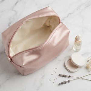 May include: A dusty rose satin cosmetic bag with a zip closure and a cream-coloured interior lining. Beside the bag sits a small clear glass dropper bottle, a round white compact case, and two sprigs of dried lavender flowers.
