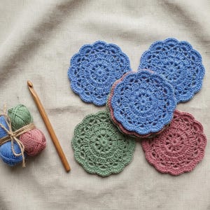 May include: A set of round, crocheted coasters in blue, green, and dusty rose colors with intricate floral patterns. A wooden crochet hook and a small bundle of matching blue, green, and dusty rose yarn tied with twine are positioned next to the coasters.