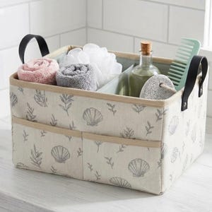 May include: A rectangular fabric storage bin with a beige base and a gray seashell and leaf pattern. The bin features two black handles, two front exterior pockets, and an internal divider containing rolled towels, a loofah, a bottle, and a comb.