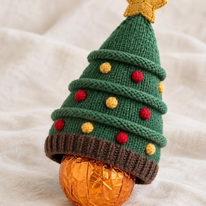 May include: A knitted green Christmas tree hat with a brown ribbed brim, topped with a yellow star. The tree is decorated with small red and yellow pom-poms and sits atop a round, orange foil-wrapped chocolate sphere.