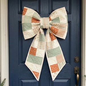 May include: A large decorative bow made of quilted fabric featuring a patchwork design. The fabric consists of squares and rectangles in muted sage green, burnt orange, and cream, with various patterns including gingham, stripes, and floral prints.