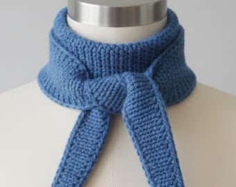 May include: A blue knitted scarf is displayed on a mannequin. The scarf is a solid light blue color and has a textured appearance. The scarf is tied in a knot at the front, with the ends hanging down. The scarf is designed to be worn around the neck.