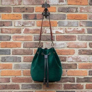 May include: A dark green velvet drawstring bag with brown leather straps and a black tassel. The bag hangs from a decorative black metal hook against a multi-colored brick wall. The bag's drawstring is tied with a knot.