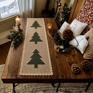 May include: A rectangular crochet table runner in a light beige yarn featuring three dark green pine tree motifs centered along the length. The runner has a delicate openwork grid pattern and a scalloped edge finish, providing a textured surface.