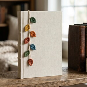 Può includere: Un libro con copertina rigida color crema dotato di un segnalibro verticale all'uncinetto attaccato al dorso. Il segnalibro è composto da un sottile cordino bianco sporco decorato con sei piccole foglie all'uncinetto in verde, giallo, arancione e blu.