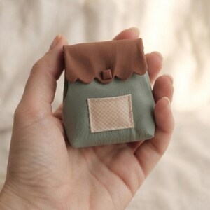 May include: A small, sage green pouch featuring a scalloped brown flap with a central square button closure. The front of the pouch displays a rectangular beige pocket with a subtle, fine grid pattern stitched onto the sage green base material.