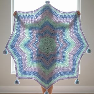 Pode incluir: Uma manta de crochê em forma de estrela com um padrão repetitivo de fios em tons pastel de lavanda, verde menta e azul claro. A manta tem um design de ponto texturizado e é finalizada com pequenas borlas de fio em cada ponta.