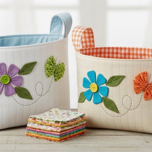 May include: Two quilted fabric storage baskets with handles. The left basket is white with a blue striped interior, a purple flower with a green button centre, and a green butterfly. The right basket is cream with an orange gingham interior, a blue flower with a yellow button centre, and an orange butterfly. A stack of colourful patterned fabric squares sits in front.
