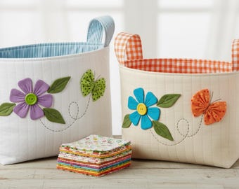 May include: Two quilted fabric storage baskets with handles. The left basket is white with a blue striped interior, a purple flower with a green button center, and a green butterfly. The right basket is cream with an orange gingham interior, a blue flower with a yellow button center, and an orange butterfly. A stack of colorful patterned fabric squares sits in front.
