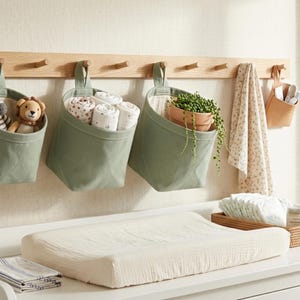May include: Three sage green fabric hanging storage baskets suspended from a light wood wall rack. The baskets contain plush animal toys, rolled white cloths, and a potted trailing plant. A patterned cloth hangs nearby on the wooden peg rail.