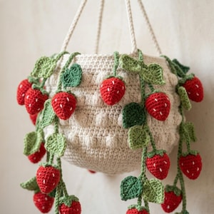 May include: A cream-colored crocheted hanging basket featuring a textured bobble stitch pattern. The basket is adorned with multiple dangling green vines, each decorated with small red crocheted strawberries with white embroidered seed details.