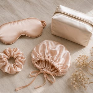 May include: A set of champagne-colored satin accessories including a sleep mask with an elastic strap, a hair scrunchie, a gathered hair bonnet with tie strings, and a rectangular zippered cosmetic pouch with a metallic zipper closure.
