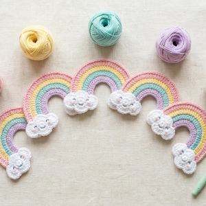 Op de afbeelding: Een reeks van vier gehaakte regenboogapplicaties met lachende witte wolken aan elk uiteinde, gerangschikt in een boog. De regenbogen hebben pastelkleurige strepen in roze, geel, taling en lavendel. Vijf bijpassende bollen garen en een haaknaald.