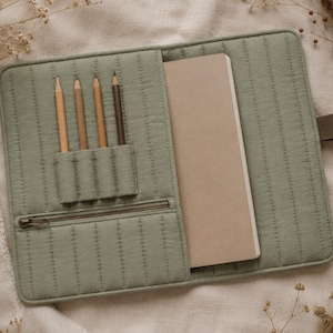 Può includere: Una copertina per taccuino in tessuto trapuntato verde salvia con motivo a cuciture verticali, un portamatite a quattro scomparti contenente quattro matite in legno, una tasca orizzontale con cerniera e una tasca interna con un taccuino beige.
