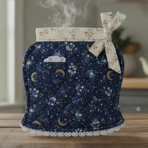 May include: A quilted navy blue tea cosy featuring a pattern of small blue flowers and gold crescent moons. The top edge has a cream-coloured floral band with a fabric bow, and the bottom edge is finished with white lace trim and a small pocket.