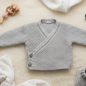 May include: A light gray long-sleeve wrap cardigan featuring a textured knit pattern, a V-neckline, and two round buttons on the side. The garment is accompanied by a wooden rattle, a sage green pacifier, a white muslin cloth, and gray knit booties.