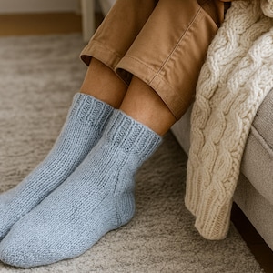 Könnte beinhalten: Ein Paar hellblaue Stricksocken. Die Socken werden mit einer beigefarbenen Hose und einer cremefarbenen Zopfstrickdecke getragen. Die Socken scheinen aus einem weichen, warmen Material zu sein, perfekt zum Entspannen zu Hause.