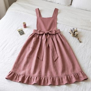 May include: A dusty rose apron featuring a square neckline, wide shoulder straps, and a gathered skirt with a ruffled hem. The apron includes a single patch pocket on the right side and a wide fabric tie at the waist that forms a large bow at the front.