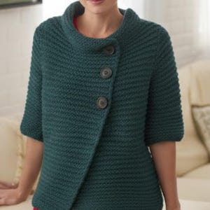 Könnte beinhalten: Eine tealgrüne Strickjacke mit asymmetrischem Knopfverschluss. Der kurzärmelige Pullover hat einen hohen, gefalteten Kragen und drei große, dunkle Knöpfe. Das Strickwerk hat ein strukturiertes Aussehen und eine lockere Passform.