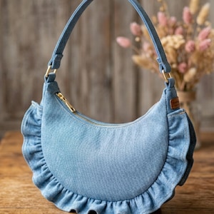 May include: A crescent-shaped denim handbag in a light blue wash featuring a ruffled trim along the bottom edge, a single shoulder strap attached with gold-tone hardware, and a gold-tone zipper closure across the top opening of the bag.