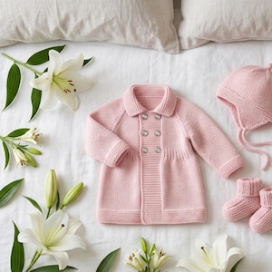 Può includere: Un completo di abbigliamento per neonati in rosa tenue, composto da un cappotto lavorato a maglia con bottoni argentati, un cappello con lacci e scarpine abbinate. Gigli bianchi e foglie verdi sono disposti su una superficie bianca.