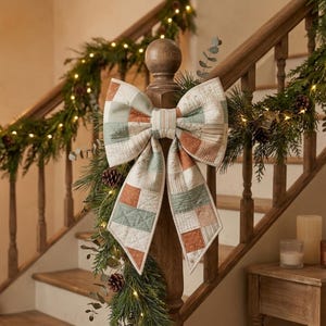 May include: A large decorative bow made of quilted fabric featuring a patchwork pattern in muted shades of sage green, terracotta orange, and cream. The bow has two wide loops and two long, pointed tails, all finished with visible stitching.