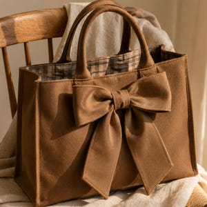Puede incluir: Un bolso tote marrón que presenta un lazo grande y prominente sujeto al panel frontal. El bolso tiene dos asas superiores resistentes y un forro interior visible con un patrón de cuadros marrones y blancos. La superficie es mate.