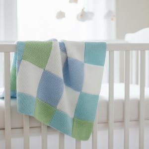 Könnte beinhalten: Eine gestrickte Decke mit einem Karomuster aus abwechselnden weißen, hellblauen und hellgrünen Quadraten. Die Decke liegt über dem weißen Gitter eines Kinderbettes und weist durchgehend ein gleichmäßiges, strukturiertes Strickmuster auf.