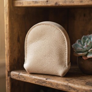 May include: A semi-circular pouch crafted from off-white, pebbled-texture material. The pouch features visible stitching along the curved edge and a structured, rounded silhouette that stands upright on its own base.