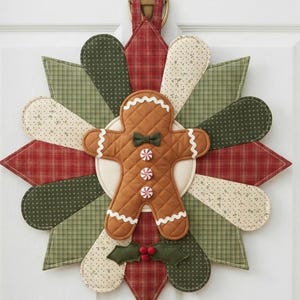 Könnte beinhalten: Ein gesteppter Wandbehang aus Stoff mit einer zentralen Lebkuchenfigur, die eine grüne Fliege und drei Pfefferminzbonbon-Knöpfe trägt. Die Figur ist von einem sternförmigen Muster aus Stoffblüten in Rot, Olivgrün und Creme umgeben.