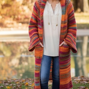 Puede incluir: Cárdigan largo y abierto con un patrón de rayas en naranja, rojo y verde azulado. La prenda de punto hecha a mano presenta mangas largas, bolsillos y un diseño hasta la rodilla. El suéter acogedor y colorido es perfecto para el otoño.