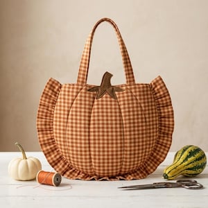 Op de afbeelding: Een pompoenvormige draagtas van oranje en wit gingham-stof met een bruine stoffen steel aan de bovenkant. De tas is voorzien van een geplooide rand, twee stoffen handvatten en verticale doorgestikte lijnen die de vorm van een pompoen nabootsen.