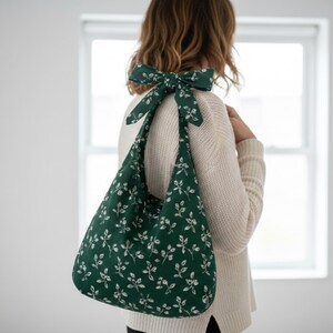 Könnte beinhalten: Eine dunkelgrüne Umhängetasche mit weißem Blumenmuster und passender Schleife. Die Tasche ist aus Stoff und hat eine gebogene Form. Die Schleife ist oben an der Tasche gebunden.