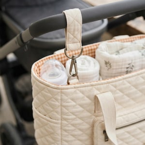 Peut inclure: Un sac fourre-tout matelassé de couleur crème avec un motif en losanges, doté d'une poche avant zippée et de deux anses. Le sac est fixé à la poignée d'une poussette par une sangle matelassée assortie et un clip en métal, contenant un biberon.
