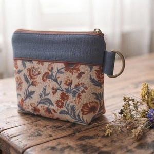 May include: A small zippered pouch featuring a cream-colored base with a floral pattern in shades of rust orange and slate blue. The top section is a solid slate blue fabric with a matching side loop and a brass-toned metal D-ring attached to the side.