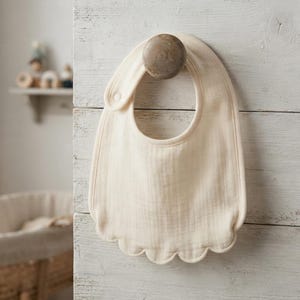 May include: A cream-colored baby bib made of soft, multi-layered muslin fabric. The bib features a rounded neckline, a single snap button closure on the side, and a decorative scalloped edge along the bottom hem, hanging from a wooden knob.