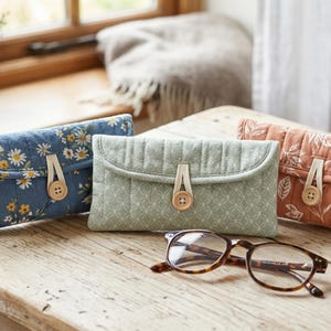 Op de afbeelding: Drie rechthoekige, doorgestikte stoffen etuis met overslagkleppen, elk beveiligd met een lichtgekleurde elastische lus en een houten knoop. De etuis hebben patronen van blauwe madeliefjes, saliegroene geometrische vormen en terracotta bladeren.