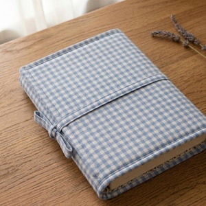 Peut inclure: Une couverture de livre rectangulaire en tissu vichy bleu clair et blanc. La couverture comporte une sangle en tissu assortie qui s'enroule autour du centre et se fixe avec un petit nœud sur le côté, enveloppant entièrement le livre.