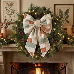 Op de afbeelding: Een groene dennenkrans versierd met kleine bruine dennenappels en warmwitte kerstverlichting. Een grote stoffen strik met een patchworkontwerp in gedempte tinten crème, saliegroen en terracotta-oranje is in het midden van de krans geplaatst.